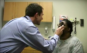A white male doctor in blue shirt examining the eye of African-American woman in grey shirt