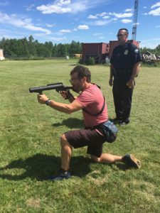 Doug wearing red shirt - kneeling and aiming a launcher with police officer standing behind him